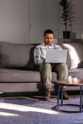 Asian young man working on laptop on gray sofa, sunlit minimalist home workspace