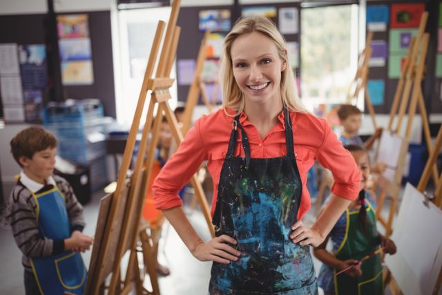 Smiling art teacher standing confidently with hands on hips in a classroom filled with young students painting on easels. Ideal for educational materials, school brochures, art class promotions, and creative learning advertisements.