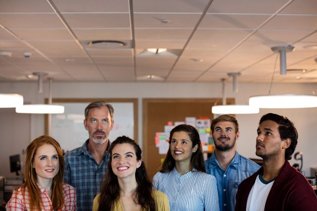 This image depicts a diverse group of creative professionals standing together in an office environment, all looking up and smiling. Ideal for use in business presentations, team-building materials, corporate websites, and promotional content highlighting teamwork, collaboration, and a positive work atmosphere.