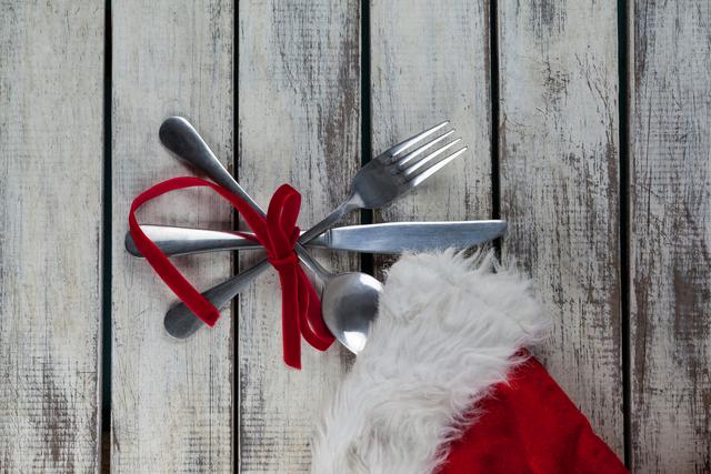 This image shows a set of cutlery tied with a red ribbon, placed on a rustic wooden table next to a Santa hat. It is perfect for holiday-themed promotions, festive dining advertisements, Christmas dinner invitations, or seasonal blog posts.