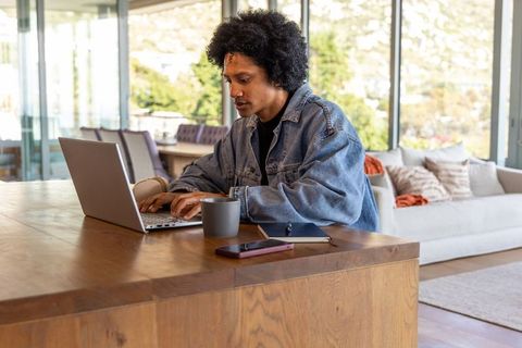 African american man working from home typing on laptop at wooden island with coffee