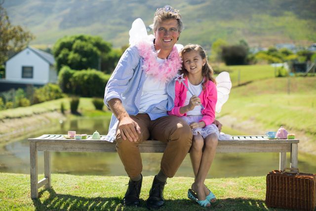 Father and daughter dressed in fairy costumes sitting on a bench outdoors, smiling and enjoying a tea party. Ideal for use in family-oriented content, parenting blogs, advertisements promoting family activities, or articles about imaginative play and bonding moments.