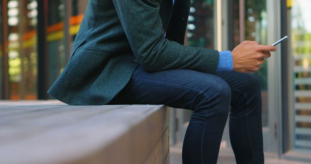 Man seated outside in a city-like area using a smartphone. Wearing casual outfit with jeans and jacket. Ideal for depicting modern urban lifestyle, technology use in public spaces, or casual fashion.