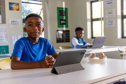 Young African American boy using tablet in classroom with classmate working on laptop