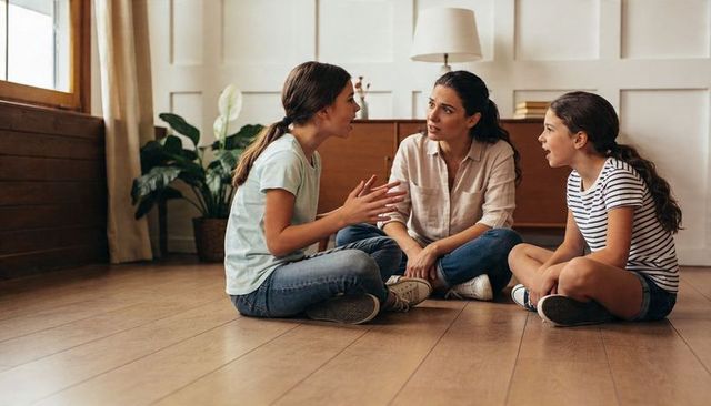 Mother listening as teen daughter talks with younger sister on living room hardwood floor