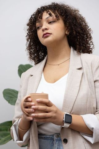 African american woman cradling ceramic mug, wearing beige blazer and smartwatch, modern portrait