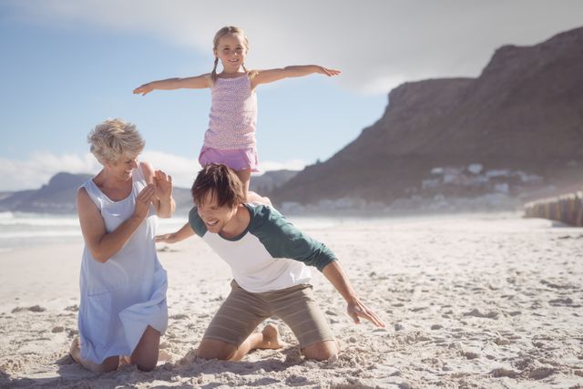 Family enjoying a sunny day at the beach. Daughter kneeling on father's back while grandmother claps. Perfect for promoting family vacations, outdoor activities, and summer fun.