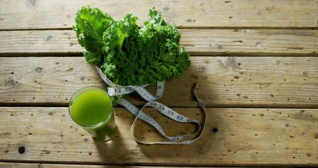 Fresh green kale leaves arranged with a glass of green juice and a measuring tape laying nearby on a rustic wooden table. Ideal for content related to healthy living, nutrition, detox plans, diet and nutrition articles, organic produce promotions, and weight loss programs. The natural and rustic setting emphasizes the freshness and organic quality of the produce.