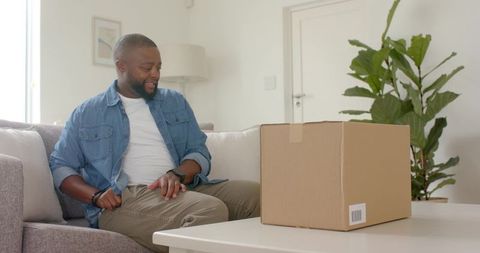 Smiling african american man sitting on sofa examining delivery box on coffee table in living room
