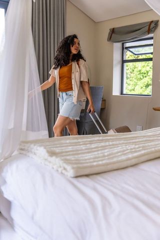 African american woman pulling suitcase into bright bedroom while parting curtain