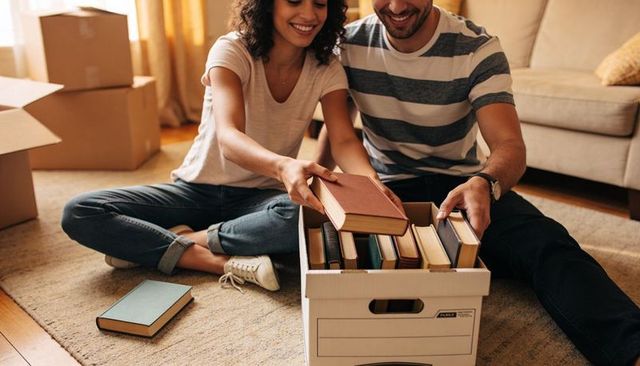 Young couple packing books into moving box on living room rug organizing personal library