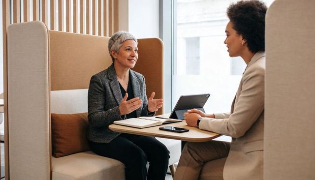 Senior executive mentoring colleague in modern office lounge with tablet and notebook