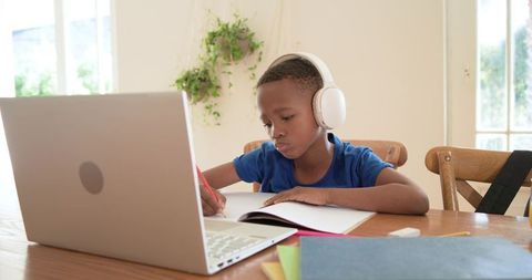 African american boy studying with headphones using laptop for remote learning at home