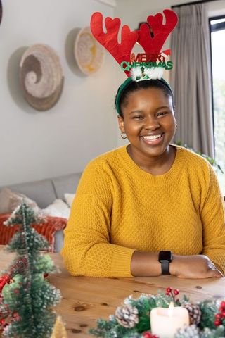 Smiling african american woman in mustard sweater with reindeer antlers decorating festive table