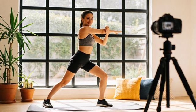 Young woman filming home workout performing wide lunge by large grid window with plants
