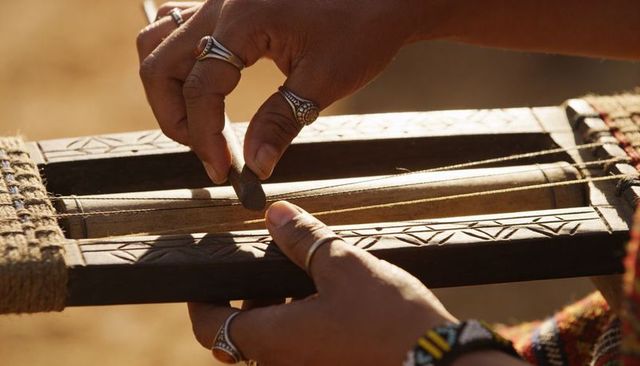 Close-up hands tuning carved wooden instrument with silver rings and woven textile in desert