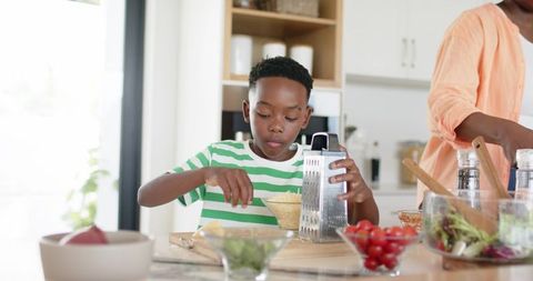African american boy grating cheese at kitchen island while parent preparing healthy meal