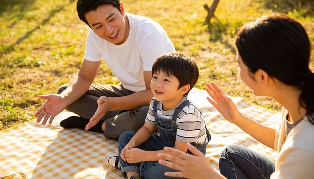 Asian family enjoying golden hour park picnic, toddler smiling on checkered blanket