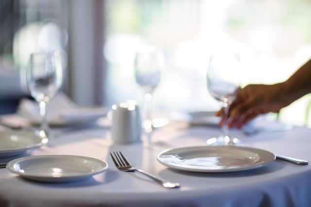 Waitress setting table in an elegant restaurant with white tablecloth, plates, glasses, and cutlery. Ideal for use in hospitality industry promotions, restaurant advertisements, dining etiquette guides, and service training materials.
