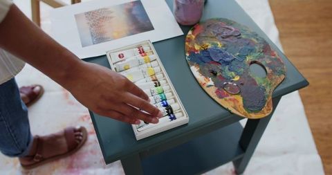 African american woman reaching for paint tubes on teal table with used palette
