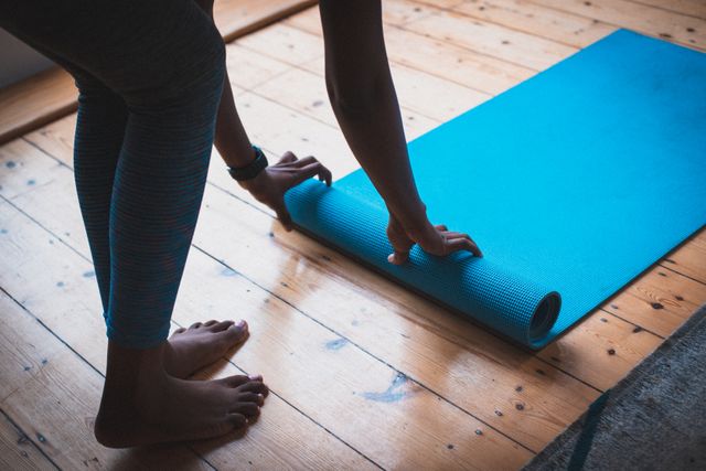 African American woman rolling up a blue yoga mat on a wooden floor. Ideal for content related to home fitness, self-care routines, quarantine activities, and healthy lifestyles. Can be used in articles, blogs, or advertisements promoting indoor workouts, yoga practices, and wellness during lockdown.