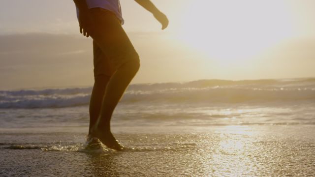 Low section of a man walking on the beach at sunset, with waves gently touching his feet. Ideal for use in lifestyle, relaxation, vacation, and nature-related projects, highlighting leisure and tranquility.