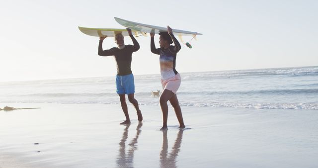 Young surfers dressed in shorts and rash guards are carrying their surfboards on a sunny beach. They are walking by the ocean with waves in the background. Ideal for advertising active lifestyles, surfing culture, and beach vacations.