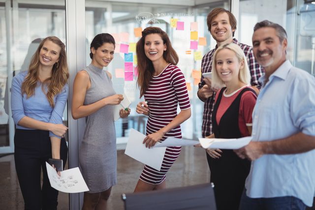 Portrait of smiling business people holding papers while standing in creative office