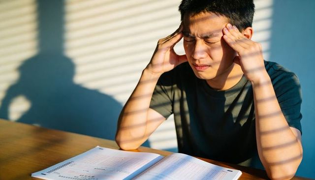 Stressed teen studying under blinds sunlight, pressing temples with exam booklet on table
