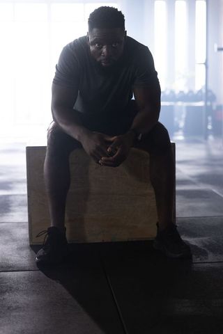 African american man resting on plyo box after intense workout in backlit industrial gym
