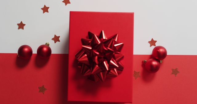 Red gift box with shiny bow surrounded by red Christmas baubles and scattered star confetti. Ideal for holiday decoration, festive background, or gift-giving themes.