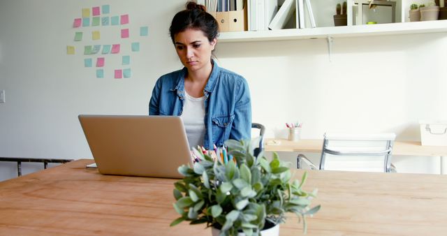 A woman working on a laptop in a modern home office. She appears focused and deep in thought. The workspace is organized with a desk, a plant, and shelves with office supplies in the background. Ideal for illustrating remote work, freelance professions, modern workspace setups, and productivity themes.