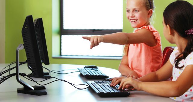 Two young girls are engaged in learning programming on desktop computers. One girl is typing while the other is pointing enthusiastically at the screen. The classroom setting with modern computers and bright environment emphasizes education and technology. Suitable for marketing technology in education, school program promotions, or articles about children's STEM education.