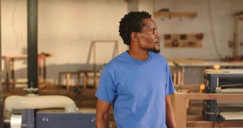 Woodworker standing in workshop wearing blue t-shirt observing woodworking machinery