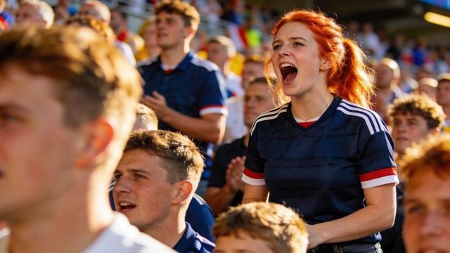 Cheering red-haired fan standing in sunlit stadium among passionate navy-jersey supporters