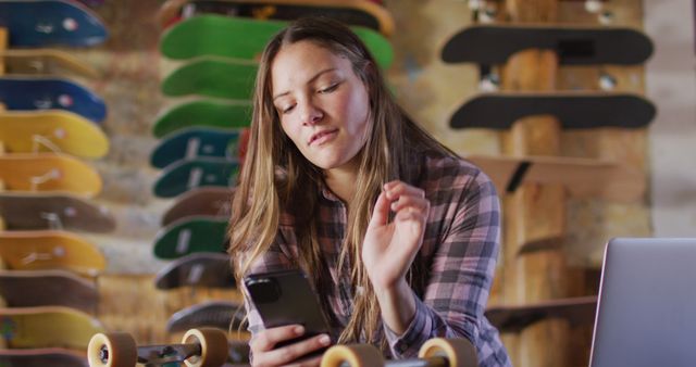 Young woman using smartphone managing skateboard shop. She wears a plaid shirt and has long hair, focusing on screen. Background shows various skateboards on wooden wall with warm lighting. Ideal for themes of small businesses, retail, modern technology in business, entrepreneurship.