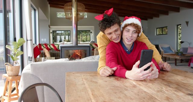Couple sitting in a cozy living room with holiday decor, including a decorated tree and garlands. The two are taking a selfie, spreading festive joy. One person is wearing a Santa hat while the other has reindeer antlers, adding a playful touch. Ideal for holiday-themed promotions, inclusivity marketing, or Christmas greeting card images.