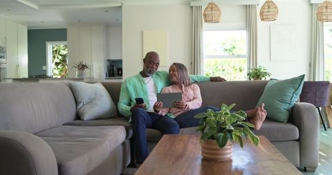 Mature african american couple relaxing on sectional sofa using tablet and smartphone