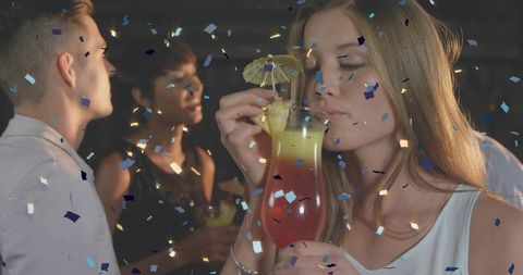 Young woman sipping tropical cocktail with umbrella amid confetti at lively night party