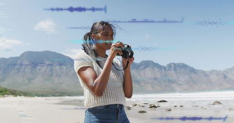 Young woman photographing coastal landscape with dslr on sunny beach and mountain backdrop