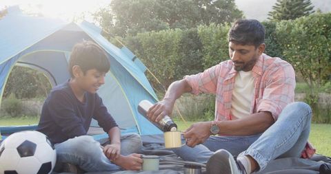 Father pouring hot drink for son while camping outdoors with blue tent sunlit family bonding