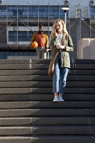 Young woman walking city steps talking on phone holding coffee, man carrying basketball