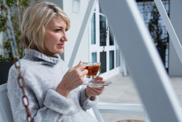 Mature woman sitting on porch, holding a cup of lemon tea, enjoying a sunny day. Ideal for use in lifestyle blogs, relaxation and wellness articles, advertisements for tea brands, and content promoting peaceful living and outdoor leisure activities.