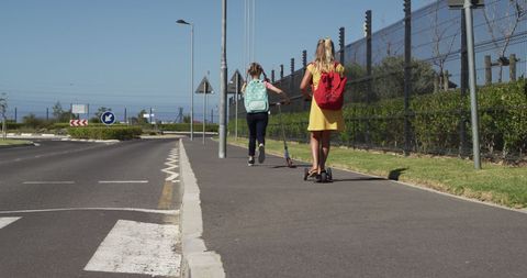 Schoolgirls Riding Scooters Along Sunny Road to School