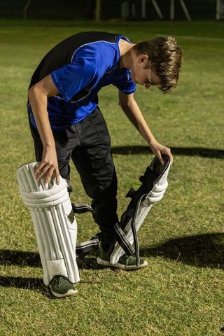 Teenage cricketer fitting white batting pads on grass under stadium lights at night