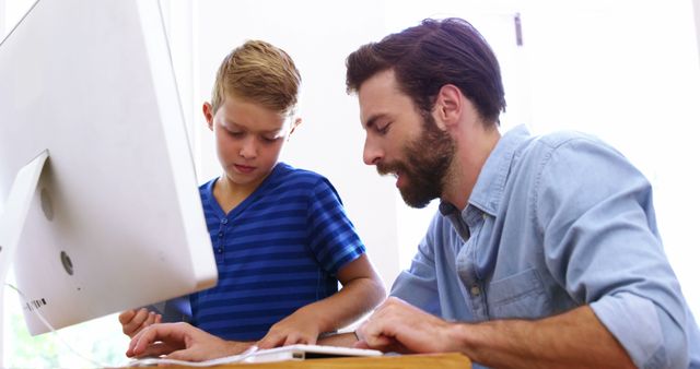 Father is helping his son with homework on a computer inside a bright room. Both look focused on the task. Image useful for themes related to technology in education, family bonding, parent involvement in learning, and home schooling.