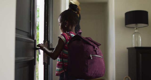 A young African American girl with a large backpack opens the front door while preparing to leave for school. The scene suggests an everyday morning routine, emphasizing themes of education, childhood, and independence. Ideal for uses in educational content, back-to-school promotions, parenting articles, or advertisements related to school supplies and children's clothing.