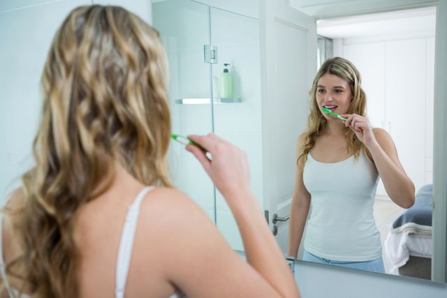 Young woman stands in front of bathroom mirror brushing her teeth. She is smiling and appears to be engaged in her daily hygiene routine. Useful for topics related to personal care, dental health, morning routines, and promoting healthy lifestyles.