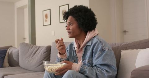 Mid-30s man sitting on couch holding popcorn bowl in denim jacket and pink hoodie watching tv