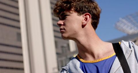 Young man looking left on sunlit urban street wearing hoodie, tank top, backpack